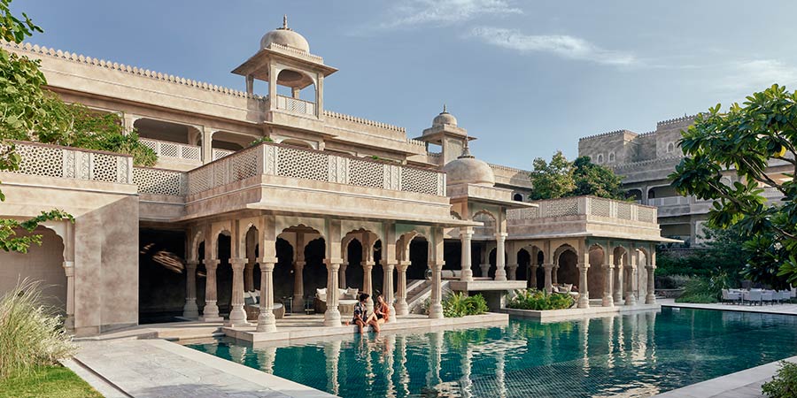 Exterior view of the Six Senses Fort Barwara hotel, a luxurious heritage hotel set within a beautifully restored 14th-century fort. The hotel features traditional Rajasthani architecture with intricate carvings, ornate windows, and a tranquil courtyard with an outdoor pool in the foreground.